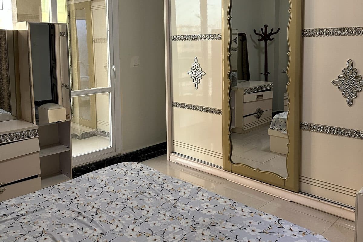 Master bedroom with a floral-covered double bed, a mirrored cream and gold sliding wardrobe, a dressing table and a glazed door letting in daylight