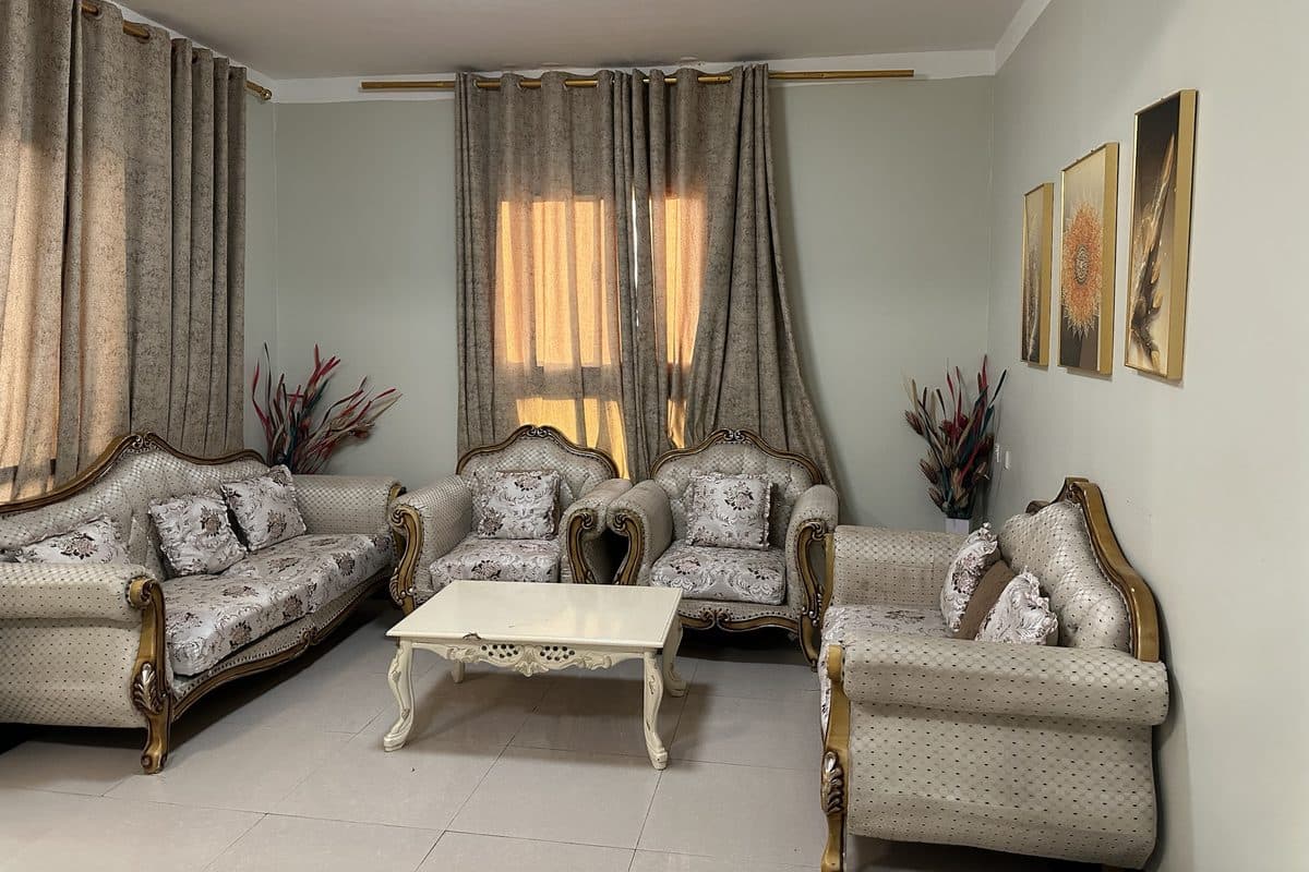 Living room with an ornate cream and gold sofa set around a white carved coffee table, sunlight through beige curtains and three framed pictures on the wall