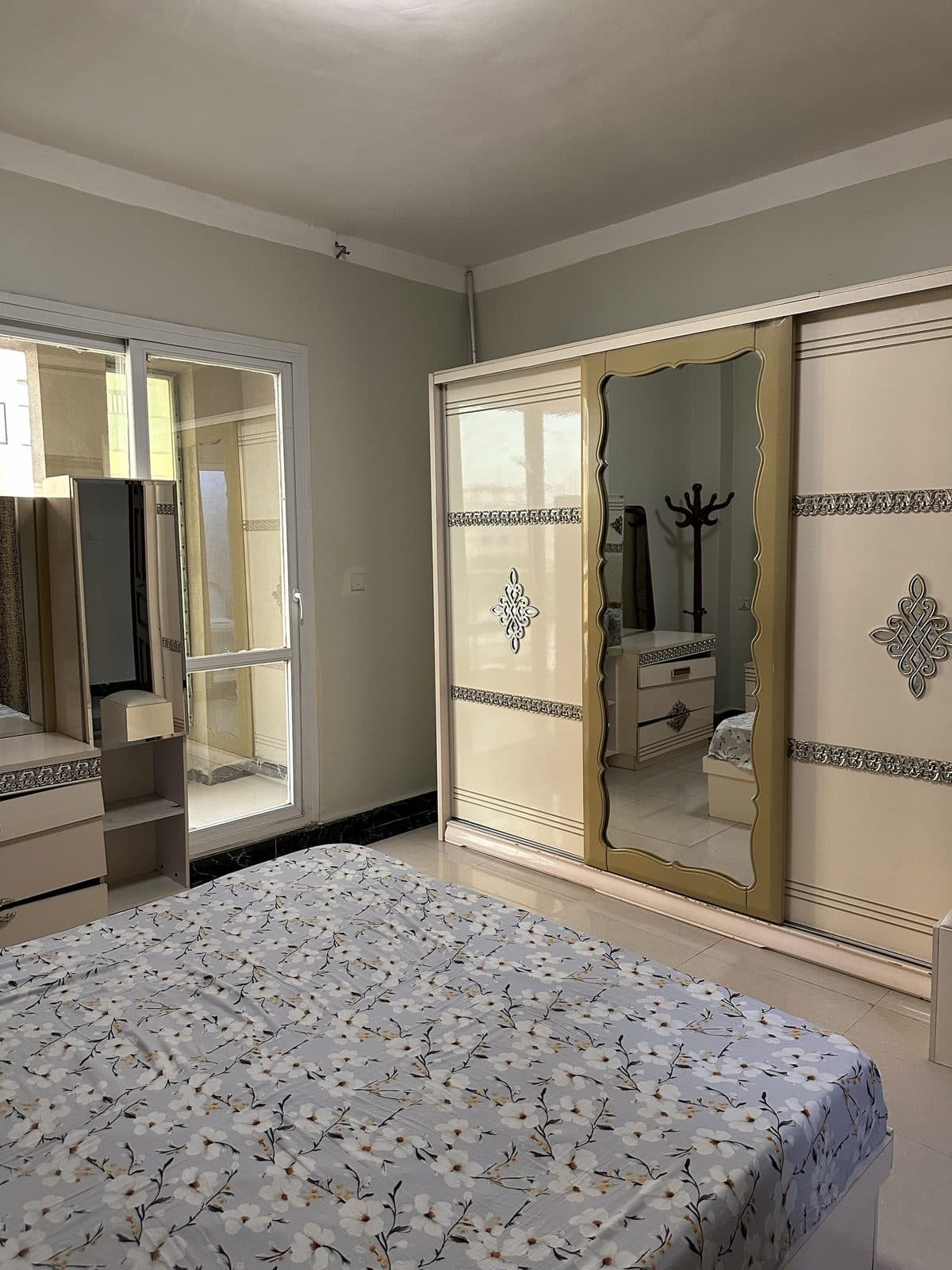 Master bedroom with a floral-covered double bed, a mirrored cream and gold sliding wardrobe, a dressing table and a glazed door letting in daylight
