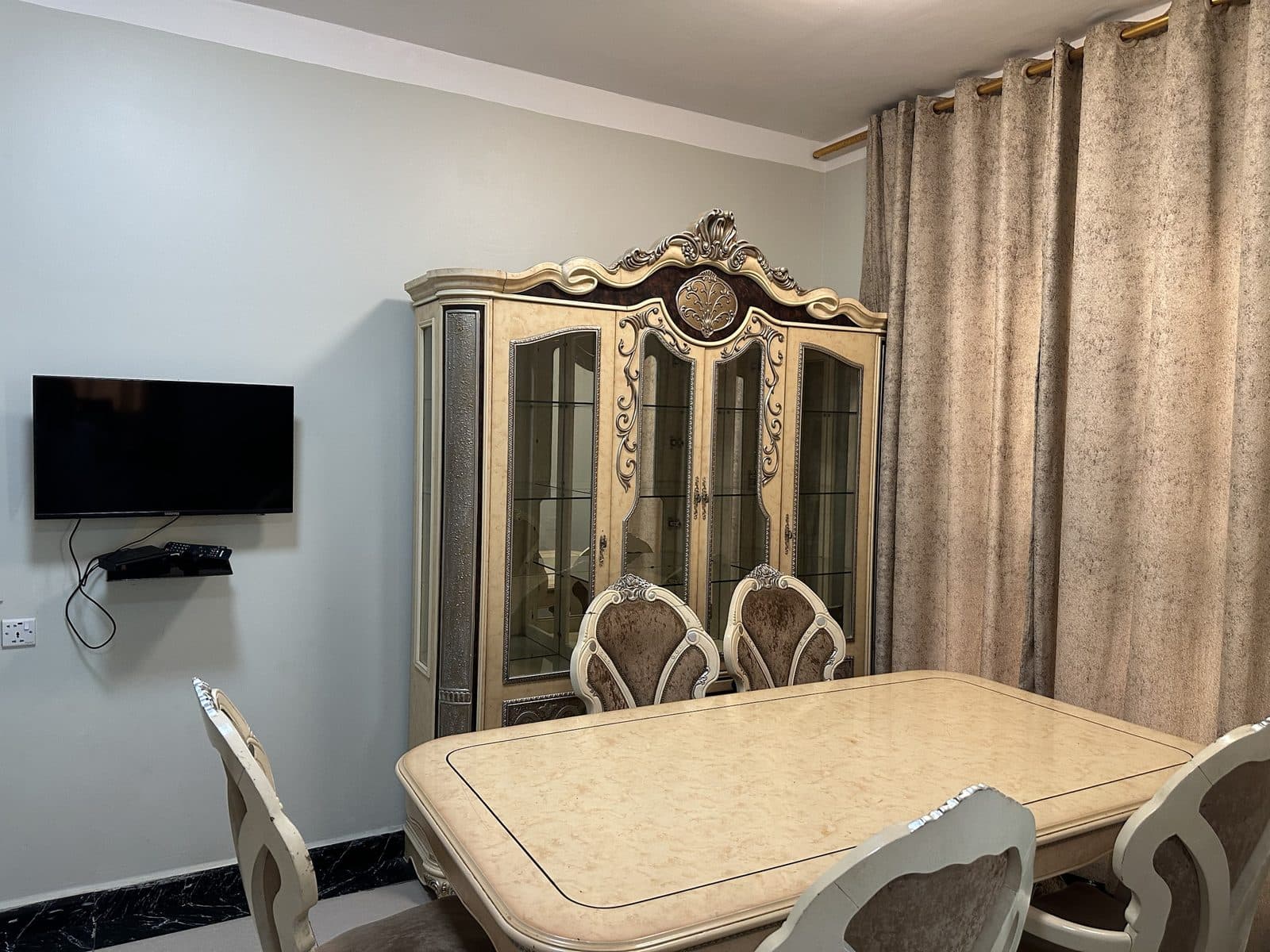 Dining room with a cream marble-effect table and carved upholstered chairs, an ornate glass-fronted display cabinet and a wall-mounted TV