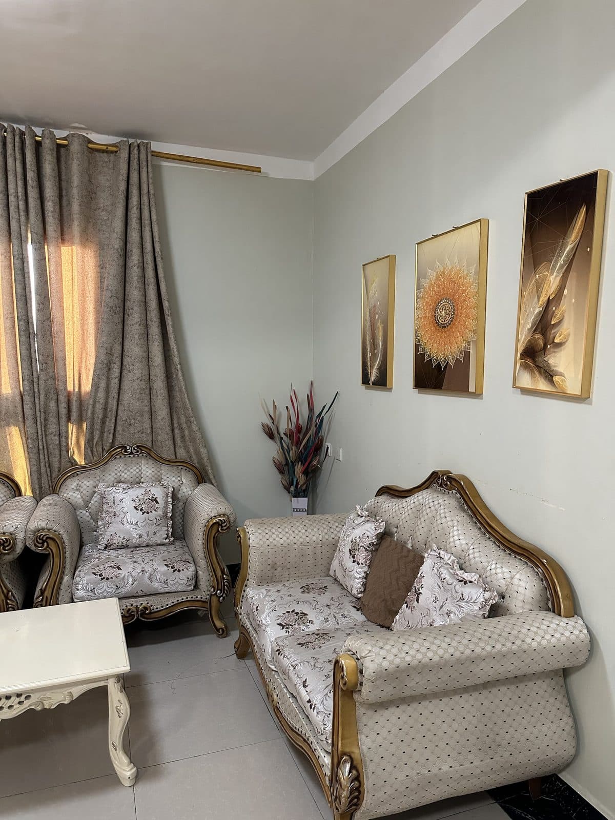 Corner of the living room with a carved armchair and two-seater sofa, floral cushions, a dried-flower arrangement and three gold-framed pictures