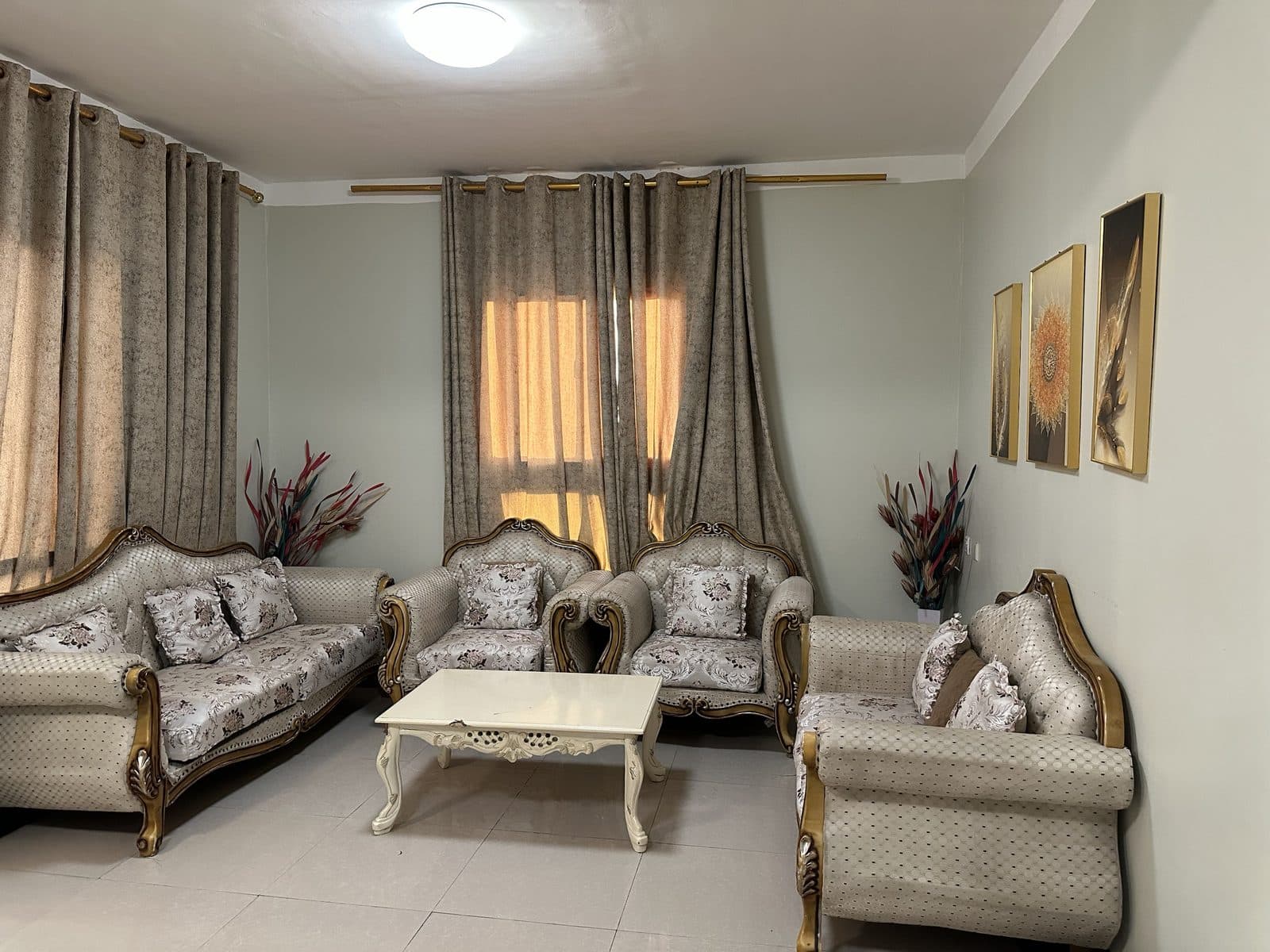 Living room with an ornate cream and gold sofa set around a white carved coffee table, sunlight through beige curtains and three framed pictures on the wall