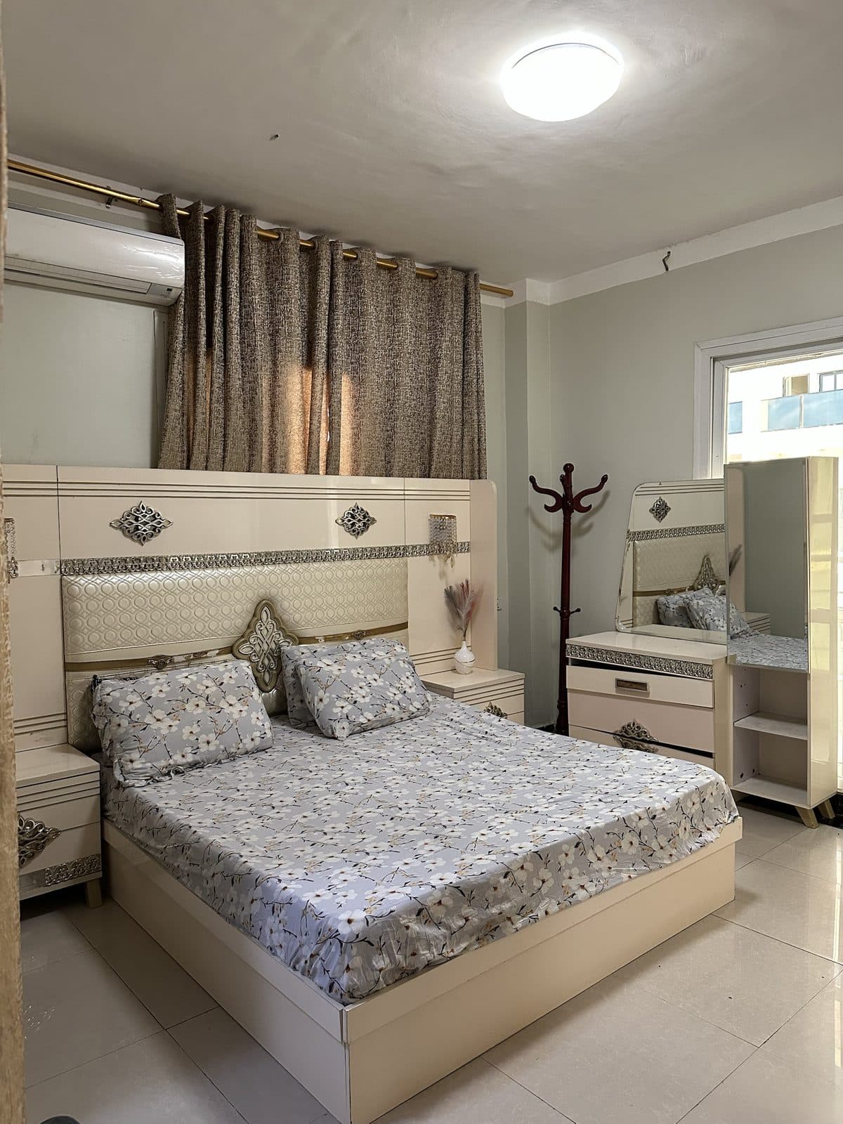 Master bedroom with a wide cream padded headboard, floral bedding, a mirrored dressing table and a coat stand by the window