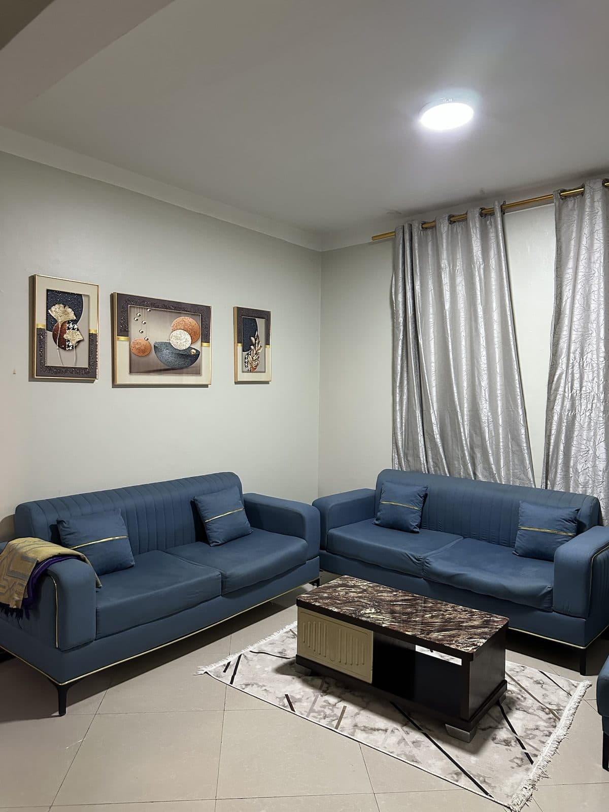 Living room with two blue velvet sofas trimmed in gold, a marble-effect coffee table, three framed artworks and silver-grey curtains