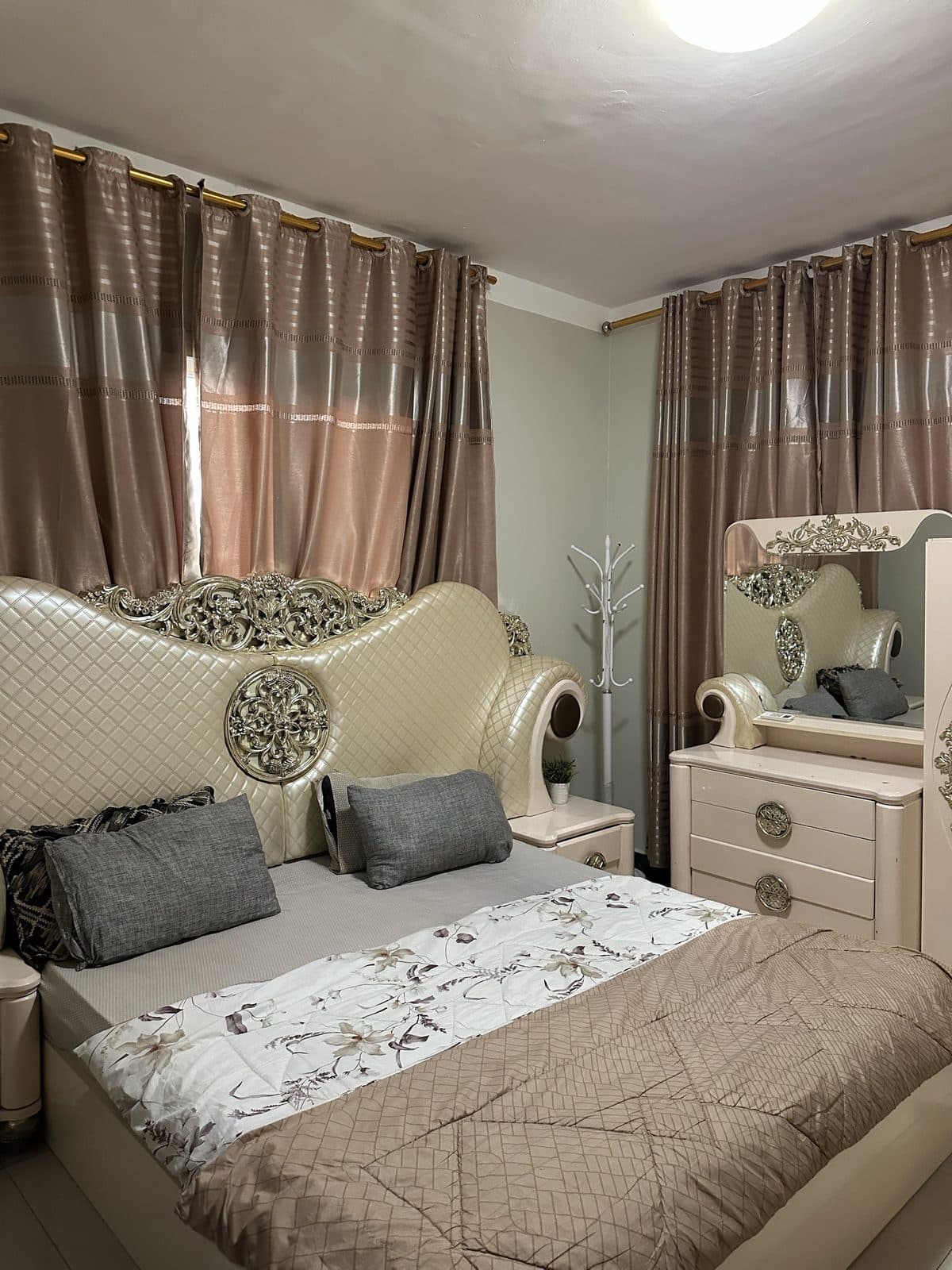 Master bedroom with a tall quilted cream headboard with gold carving, grey cushions, a floral throw and a mirrored dressing table
